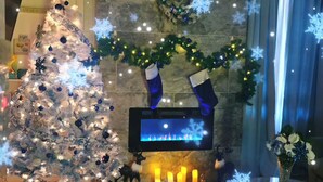 Ein festlich geschmückter Weihnachtsbaum mit Ornamenten und Lichterketten, ein Kamin mit Kerzen, darüber hängende Weihnachtssocken und ein Fernseher an der Wand.