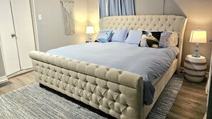 Ein Schlafzimmer mit einem gebürsteten Kopfteil, einem großen Bett mit blauer Bettwäsche, zwei Nachttischlampen, einer Wandweberei aus Makramee und einer Tür mit weißem Rahmen.