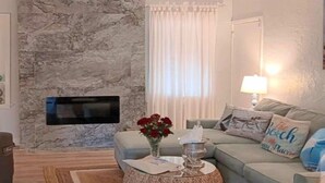 Ein Wohnzimmer mit Steinwand, einem flachbildfernseher darüber, einem runden Couchtisch mit einer Vase roter Blumen, einem hellblauen Sofa mit Zierkissen und einer Lampe auf einem Beistelltisch.