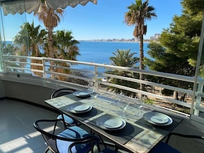 A balcony with a table set for dining, overlooking a coastal cityscape with palm trees and a clear blue sky.