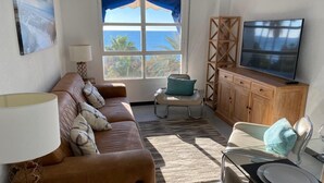 Ein Wohnzimmer mit einer Couch, einem Esstisch mit Stühlen, einem Fernseher und Meerblick durch ein Fenster.