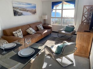 A living room with a brown leather sofa, a glass dining table set with plates and wine glasses, a blue chair, and a view of the ocean through a large window.