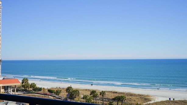 Vista de una playa con cielo despejado, arena y algunas personas caminando.