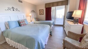 Habitación de hotel con dos camas, ventilador de techo, televisión, silla de mimbre y balcón con vistas.