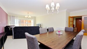 Un coin repas avec une table en bois, six chaises, un canapé noir, un lustre et une vue sur un immeuble à travers de grandes fenêtres.