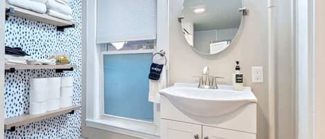 A bathroom with a white sink, a round mirror, and shelves with towels and toiletries.