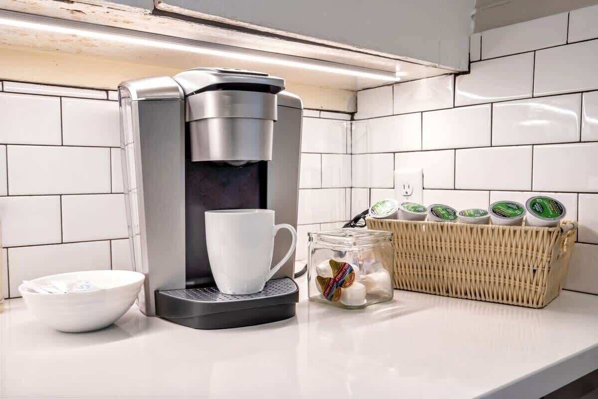 Een keuken met een koffiezetapparaat, een witte mok, een kom en een mandje theezakjes.