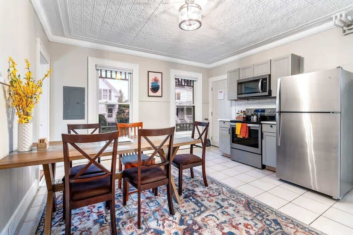 A modern kitchen with a dining area, featuring a wooden table and chairs, a patterned rug, and stainless steel appliances.