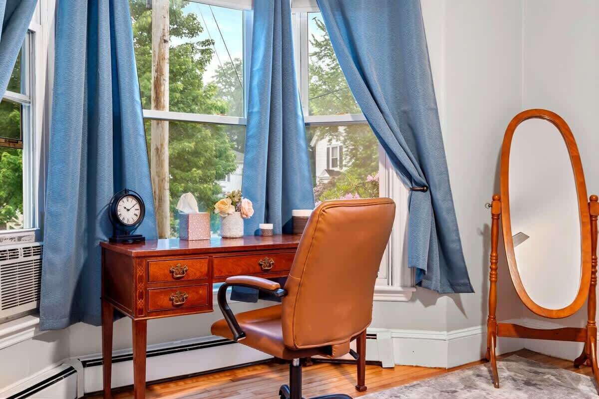 Una habitación con escritorio de madera, una silla, un reloj, un jarrón con flores, un espejo sobre un pedestal y una ventana con cortinas azules.