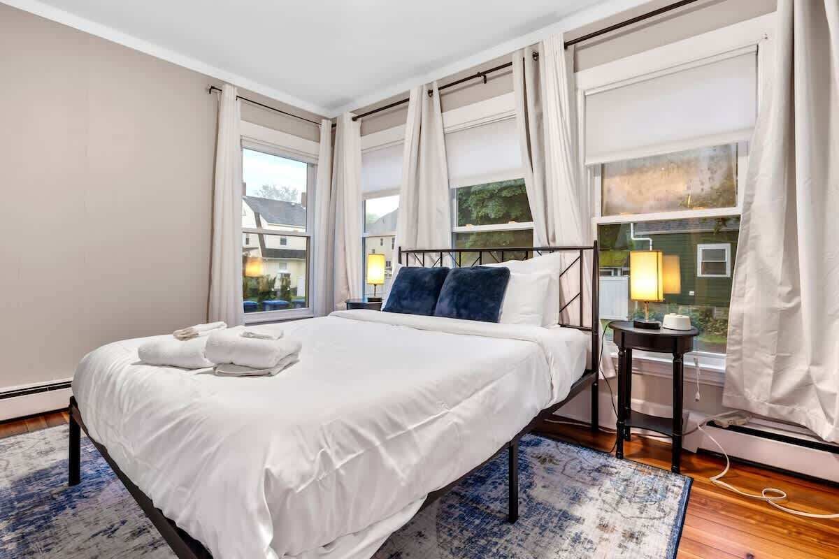 A hotel room with a large bed, a bedside table with a lamp, and a view of the outside through large windows.
