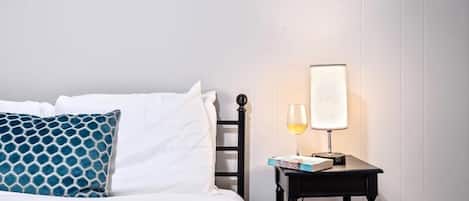 A neatly made bed with white and blue patterned pillows, a bedside table with a lamp and a glass of wine, and a framed picture on the wall.