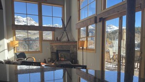 A room with large windows offering a view of snow-capped mountains. There is a fireplace with a stone surround and a dining area with a table and chairs. - Comfortable large mountain home with stunning views, hot tub and pool table! (Telluride)