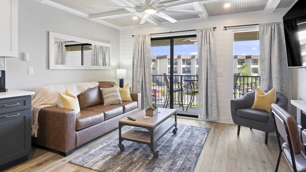 Un salon avec un canapé en cuir marron, une table basse en bois et un fauteuil gris. On y trouve un ventilateur de plafond, une grande fenêtre avec des rideaux et une vue sur un balcon.