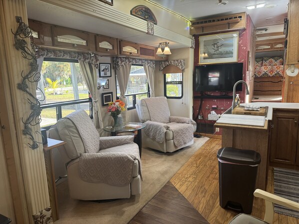 A cozy RV interior with two cushioned chairs, a small table with a vase of flowers, a television, and a compact kitchenette.