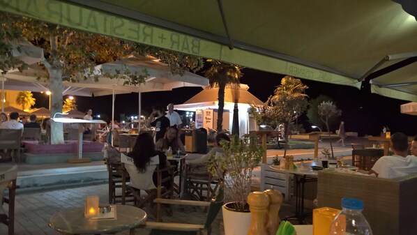 An outdoor dining area with tables and chairs, a food counter, and umbrellas providing shade.