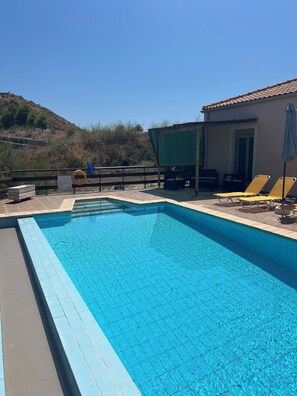 Ein Schwimmbecken mit gefliesten Rändern, ein überdachter Sitzbereich mit Stühlen und ein Gebäude mit einem Flachdach im Hintergrund.