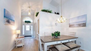 A dining area with a wooden table, white chairs, and a bench. There are two pendant lights, a small table with a lamp, and a plant on the wall.