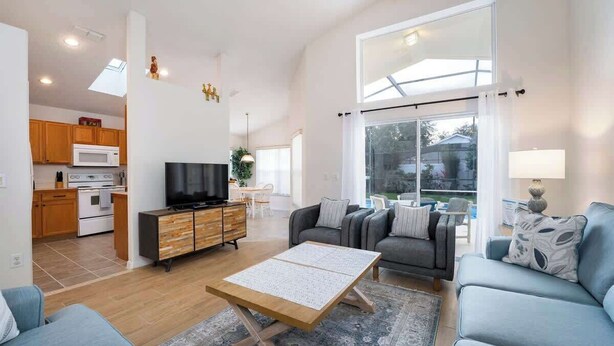 A spacious living room with a sofa, coffee table, and a television. There is a kitchen area with wooden cabinets and a microwave. The room has large windows and a view of the outdoors.