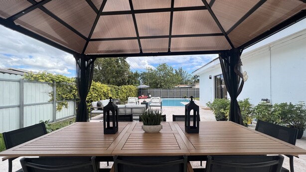 En overdækket terrasse med et træbord og stole, en potteplante og udsigt over en baghave med en pool og havemøbler.