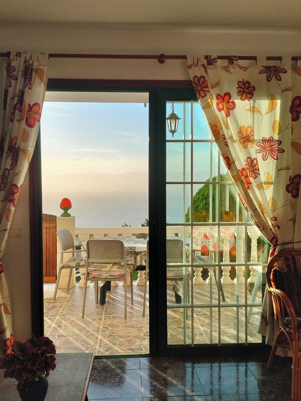 A room with a view of a terrace, a wooden door, a table with chairs, and a vase with flowers.