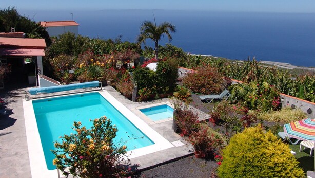 A swimming pool with a view of the ocean, surrounded by lush greenery and a small building with a red roof. - Apartments Finca House Garden (Tijarafe. La Palma)