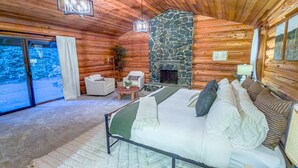 A log cabin bedroom with a stone fireplace, a bed with pillows, a chair, and a view through a glass door.