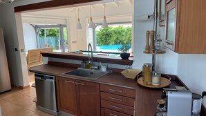 A modern kitchen with a built-in dishwasher, wooden cabinets, and a view of a swimming pool. - Villa (Baie-Mahault)