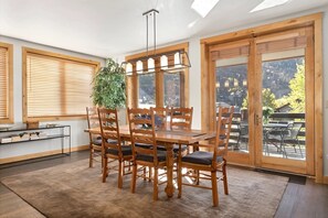 Un espace repas comprenant une table et des chaises en bois, un luminaire suspendu et de grandes fenêtres équipées de stores.