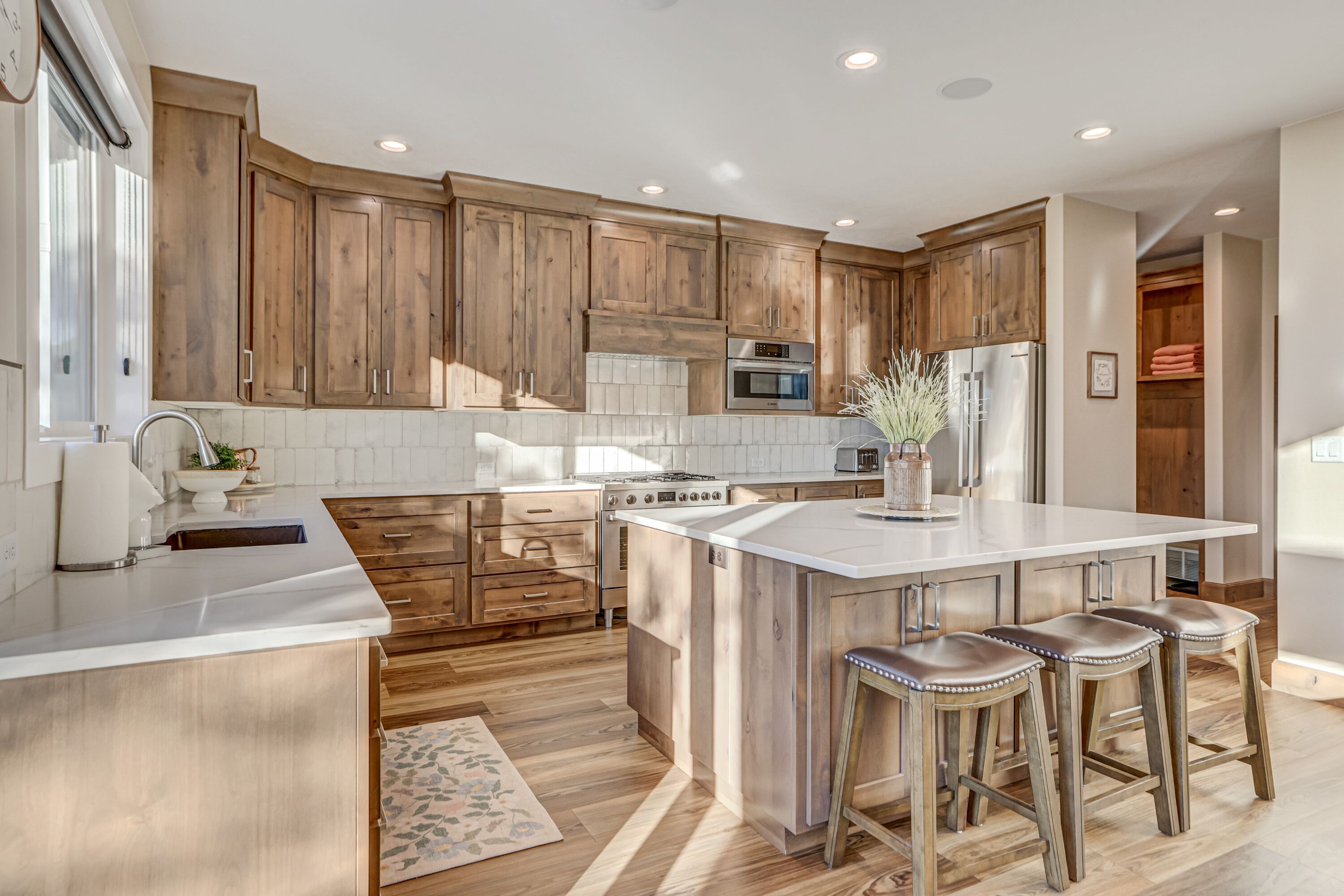 A modern kitchen with wooden cabinets, a central island with stools, stainless steel appliances, and a patterned rug.