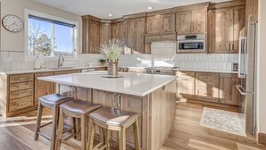 Une cuisine spacieuse avec des meubles en bois, un grand îlot central doté de tabourets, une horloge au mur et une fenêtre laissant entrer la lumière naturelle.