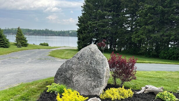 A landscaped garden with a large rock centerpiece, surrounded by various shrubs and a view of a lake and forest in the background.