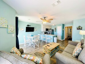 A spacious living room with a sofa, a coffee table, and a TV. There is a kitchen area with a dining table and chairs. - Grand Beach 315 - Beach View - Great location in the heart of Gulf Shores! (Gulf Shores)