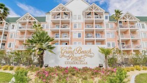 Un edificio de varios pisos con balcones, un cartel que dice "The Grand Beach Resort Condominium" y un jardín ajardinado con flores y palmeras.