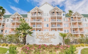 Ein mehrgeschossiges Gebäude mit Balkonen, einer Beschilderung mit dem Aufdruck "The Grand Beach Resort Condominium" und einem angelegten Garten mit Blumen und Palmen.