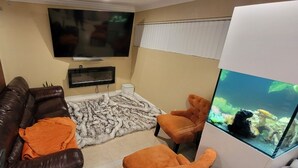 Un salón con un sofá de cuero marrón, sillones naranja, un televisor de pantalla plana, un acuario y una chimenea.