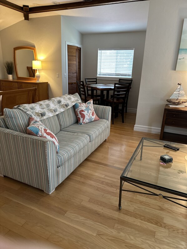 Una sala de estar con un sofá a rayas, una mesa de centro de vidrio, piso de madera, una zona de comedor con mesa y sillas, y una ventana con persianas.