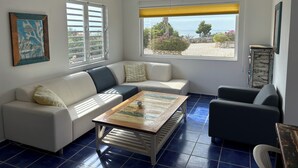 Un salon avec un canapé blanc, un fauteuil bleu, une table basse et une vue sur la plage.