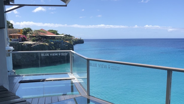 Une terrasse dotée d’une barrière en verre, offrant une vue sur des eaux turquoise et une ville côtière.