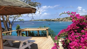 Une terrasse en bois avec une table de pique-nique, offrant une vue sur la côte, avec des bateaux et des fleurs de bougainvillier.