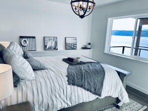A bedroom with a large bed, a chandelier, artwork on the wall, and a view of the water through a window.