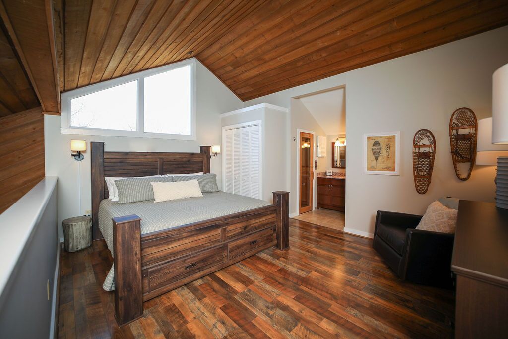 Un dormitorio con techo de madera, una cama grande con cabecera, suelo de madera, una ventana y una puerta que da a un baño.