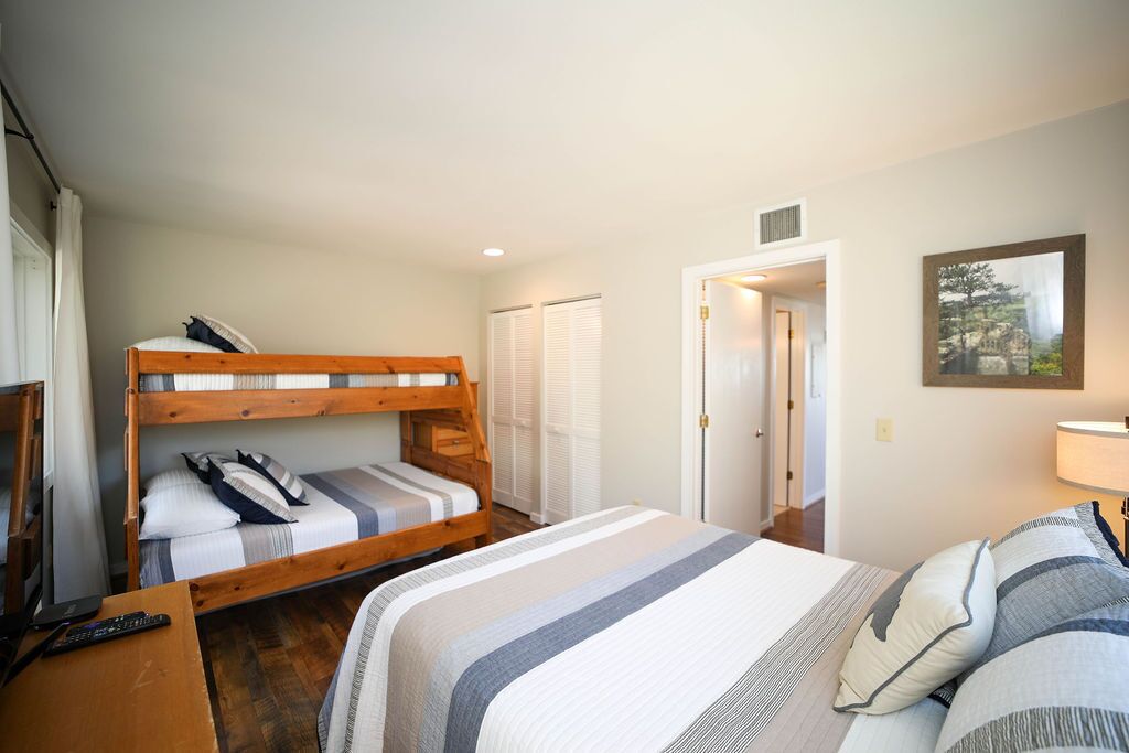 A bedroom with a bunk bed, a single bed, a desk with a lamp, and a door leading to another room.