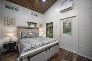 Un dormitorio con techo de madera, paredes blancas, una cama grande con ropa de cama estampada, dos mesitas de noche con lámparas y una puerta con ventana.