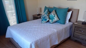 Un lit bien fait, recouvert d’une courtepointe blanche matelassée et agrémenté de coussins décoratifs turquoise, une table de chevet en bois avec une lampe, et une vue sur l’extérieur par une fenêtre ornée de rideaux bleus.