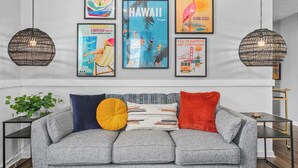 Un salon moderne avec un canapé gris, des coussins décoratifs et des œuvres d’art accrochées au mur représentant différentes destinations de voyage.