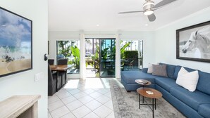 Un salon moderne avec un canapé bleu, une table basse et un ventilateur de plafond. Une grande fenêtre offre une vue sur les palmiers, et il y a un balcon.