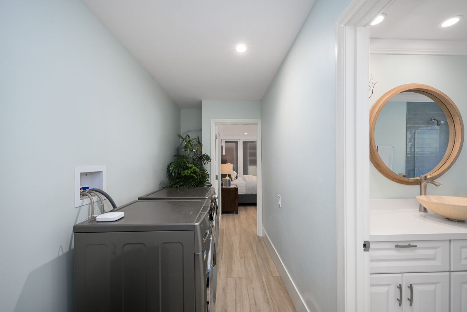Un couloir étroit, avec un coin buanderie sur la gauche, une chambre à coucher avec un lit et une table de chevet au centre, et une salle de bain avec un lavabo et un miroir sur la droite.