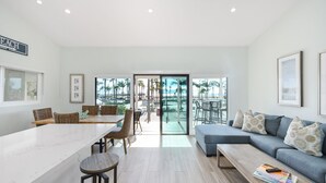 Ein modernes Wohnzimmer mit großem Fenster, einem Essbereich mit Korbstühlen und einer Couch mit gemusterten Kissen.