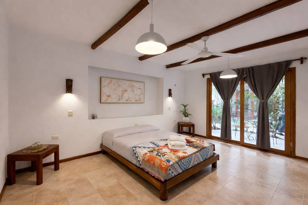 A bedroom with a bed, a ceiling fan, a window with curtains, and a wall-mounted light fixture.