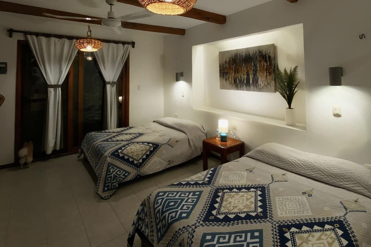 A hotel room with two beds, a large window with curtains, a ceiling fan, a wall-mounted lamp, and a painting.